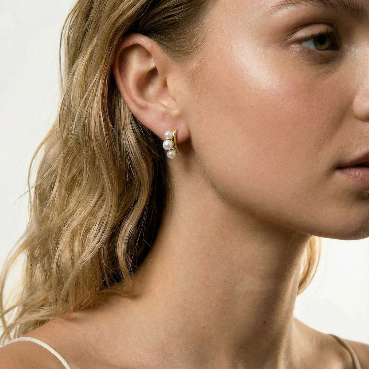 14K gold-plated curved stud earrings with three white freshwater pearls aligned vertically on cream background