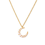 Gold crescent moon pendant with graduated white pearls on delicate chain against cream background