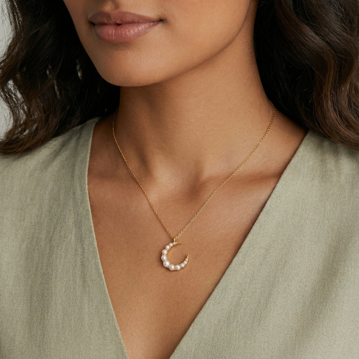 Gold crescent moon pendant with graduated white pearls on delicate chain against cream background